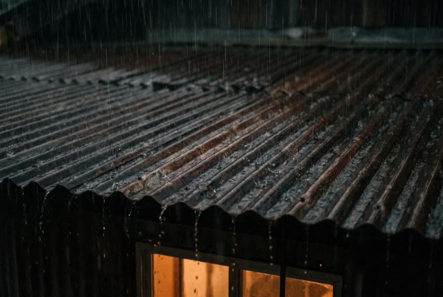 Steady rain falling on a dark tin roof at night, a warm window glowing below the eaves